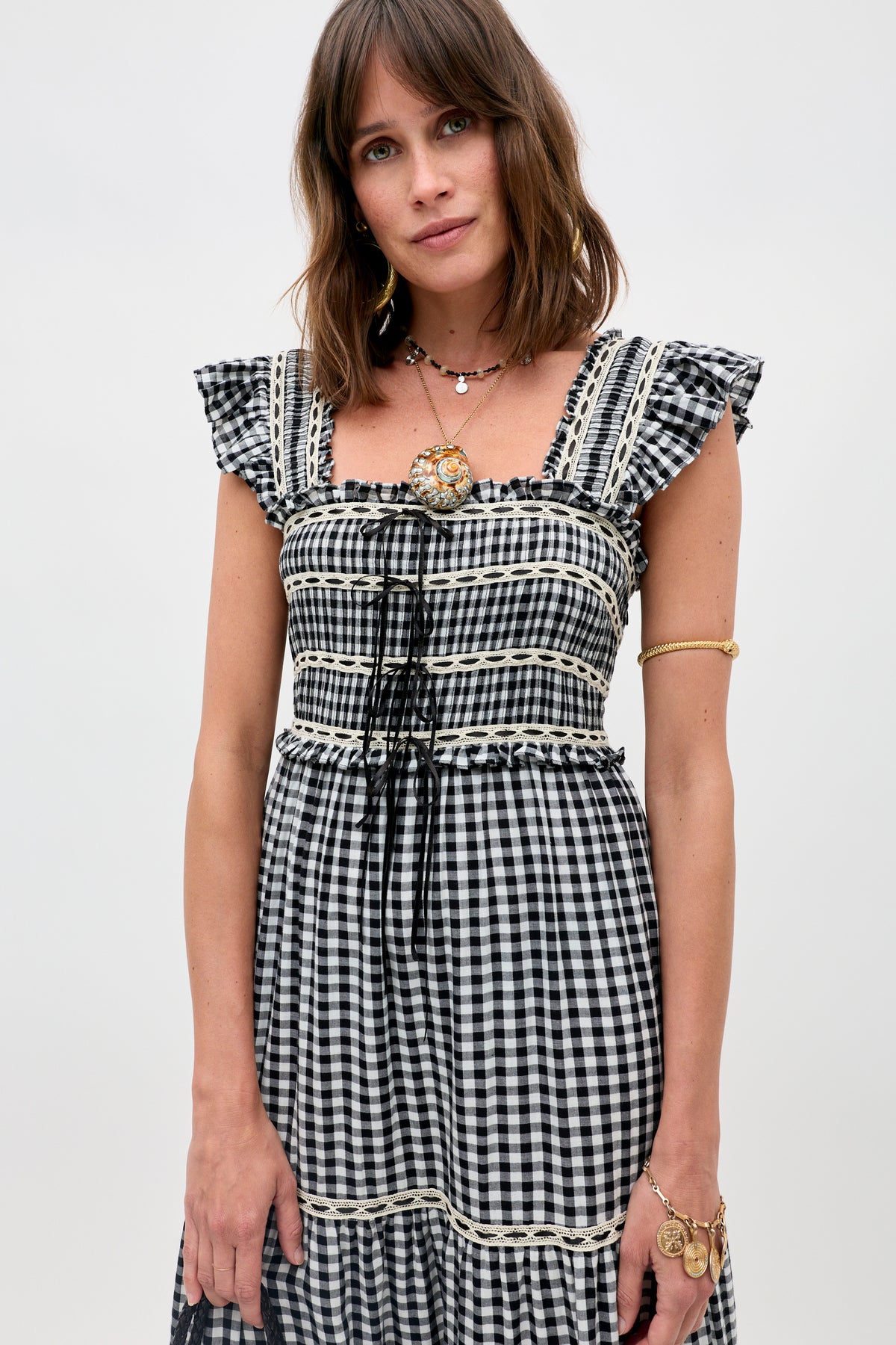Woman in the Mikah - Silver dress, a black and white gingham style with ruffled sleeves, accessorized with gold jewelry and a brooch, standing against a plain light background.