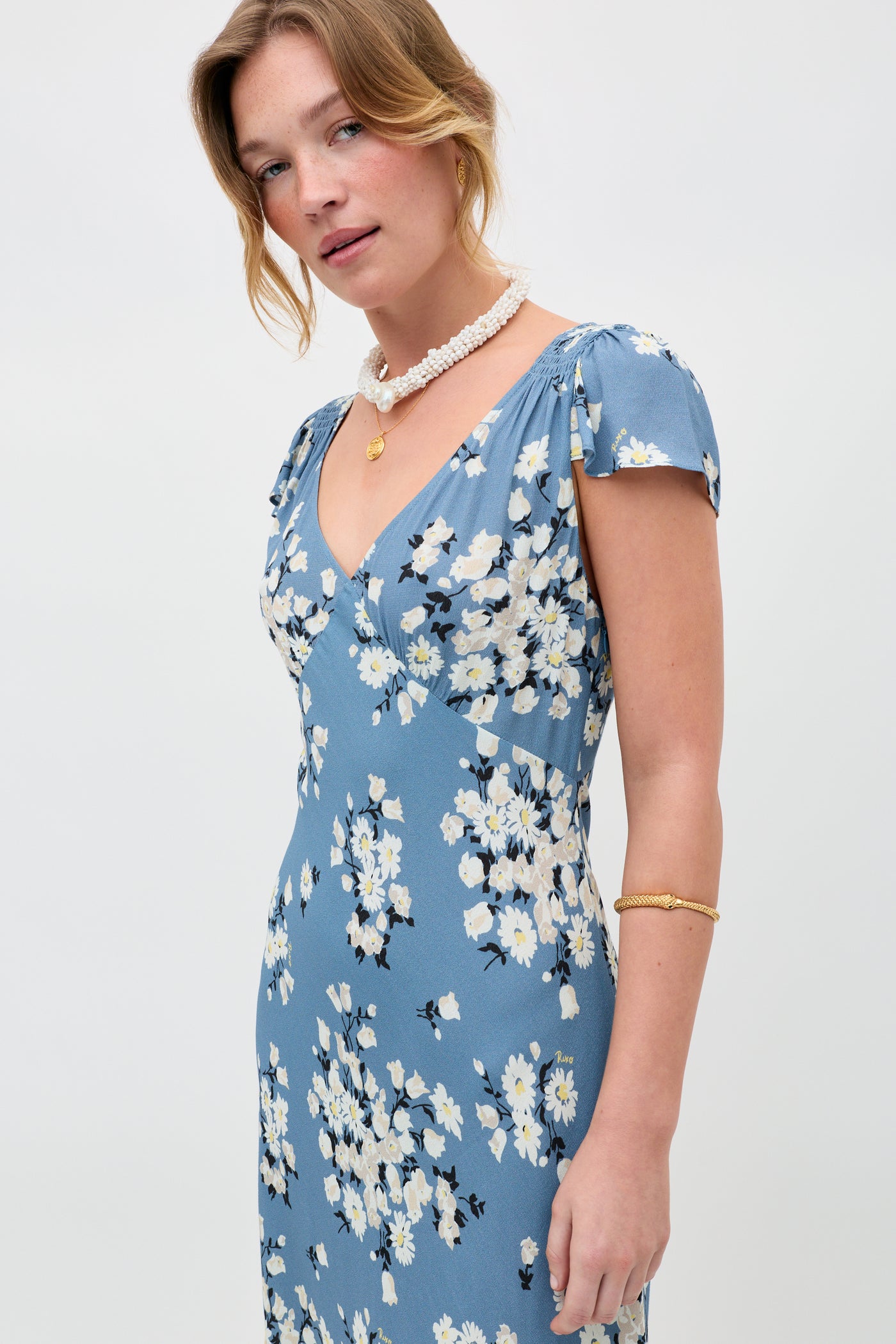 A woman models the Cathy - Amelia Bouquet Blue dress, featuring a blue floral print and short sleeves, paired with a white beaded necklace and a gold bracelet, standing against a plain white background.