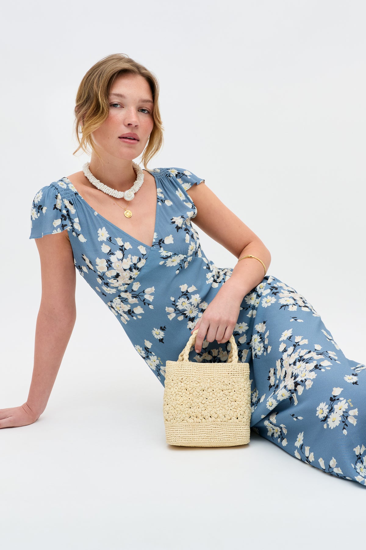 Wearing the Cathy - Amelia Bouquet Blue dress, a woman sits against a white background with a small straw handbag, white beaded necklace, gold pendant and bangle, her hair styled loosely.