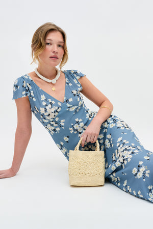 Wearing the Cathy - Amelia Bouquet Blue dress, a woman sits against a white background with a small straw handbag, white beaded necklace, gold pendant and bangle, her hair styled loosely.