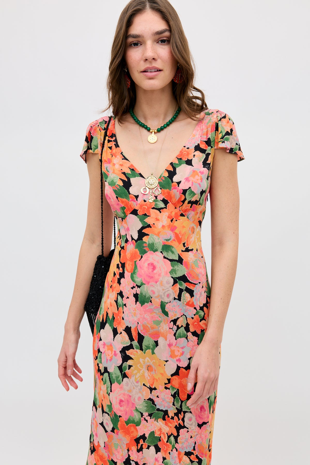 A woman with long brown hair wears a colorful floral dress and layered necklaces, including the Ischia - Green necklace with malachite crystals. She holds a black bag and stands against a plain light background.