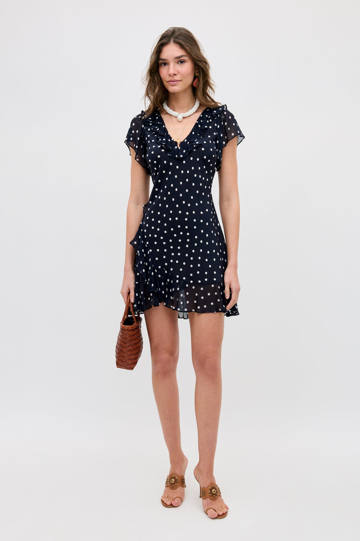 A woman stands against a plain background wearing the Tally - Layla Dot Navy short, ruffled dress with white polka dots, tan heeled sandals, a white necklace, and holding a brown woven handbag.