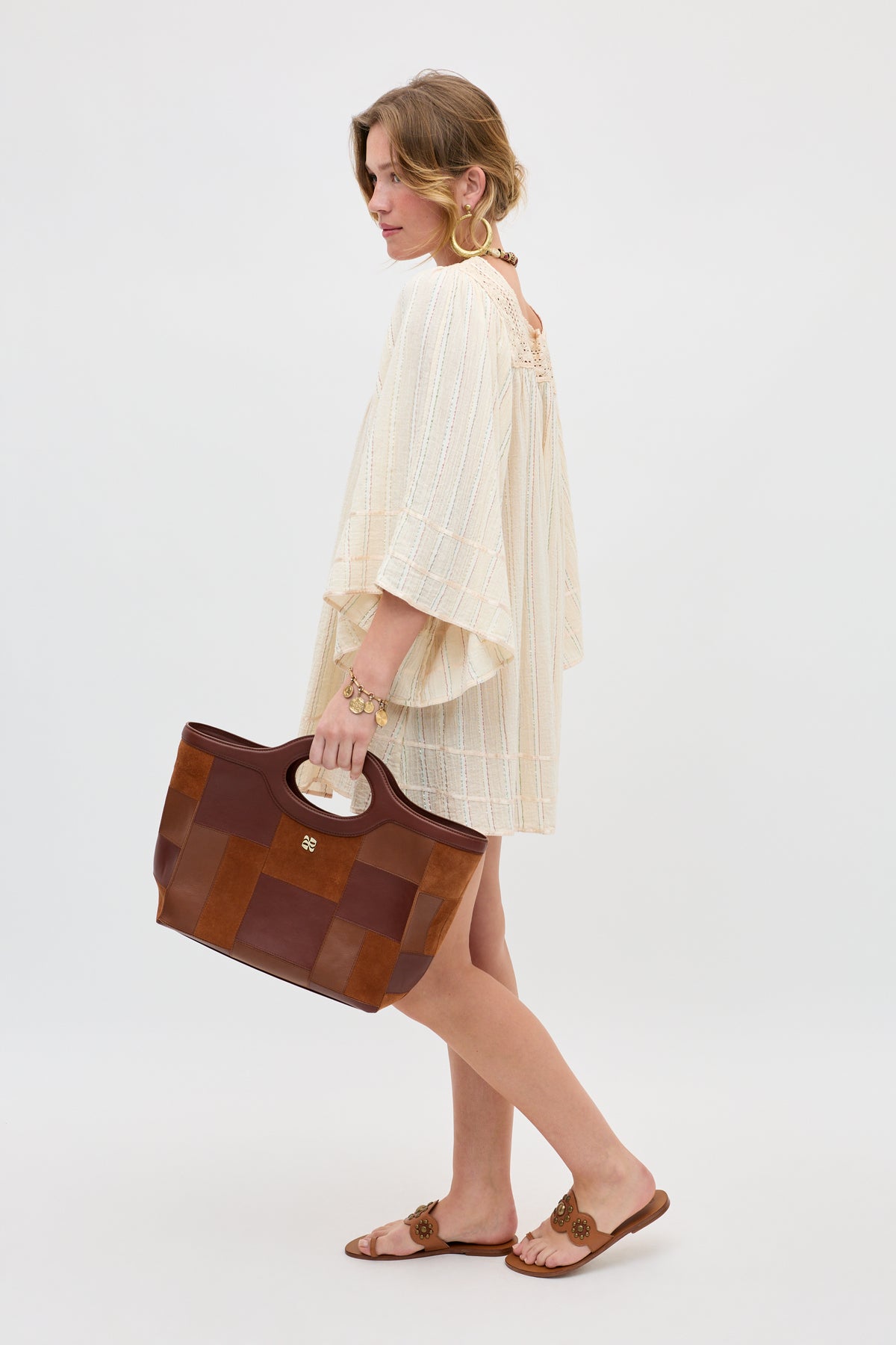 A woman stands sideways against a plain white background, wearing the Estele - Cream dress. She holds a large brown tote bag, pairs tan sandals and gold bracelets with her look, and has her hair loosely tied back.