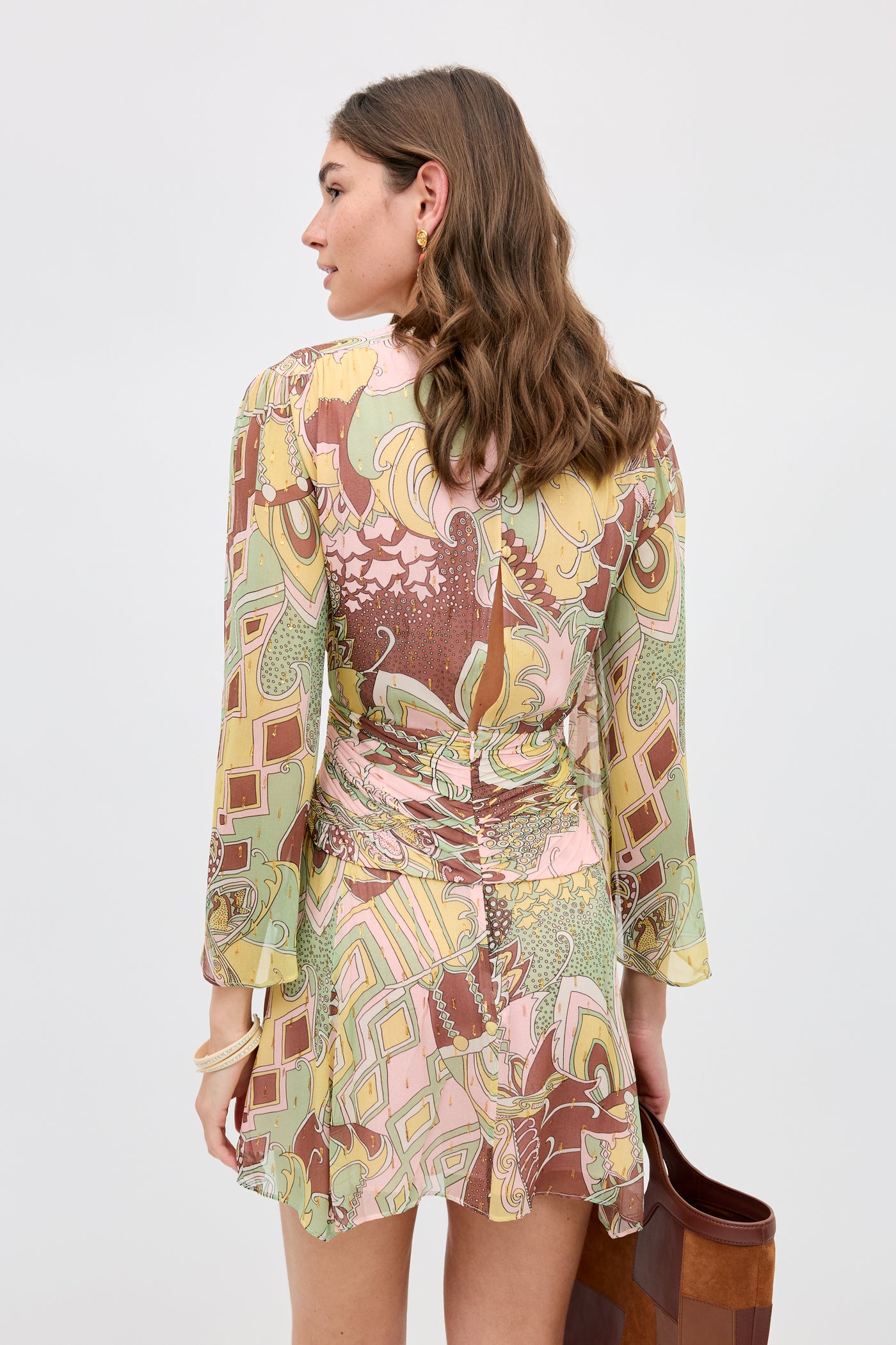 A woman with wavy brown hair is seen from behind wearing the Clove - Bianca Abstract Green dress with long sleeves and a ruffled hem, holding a brown bag against a plain light background.