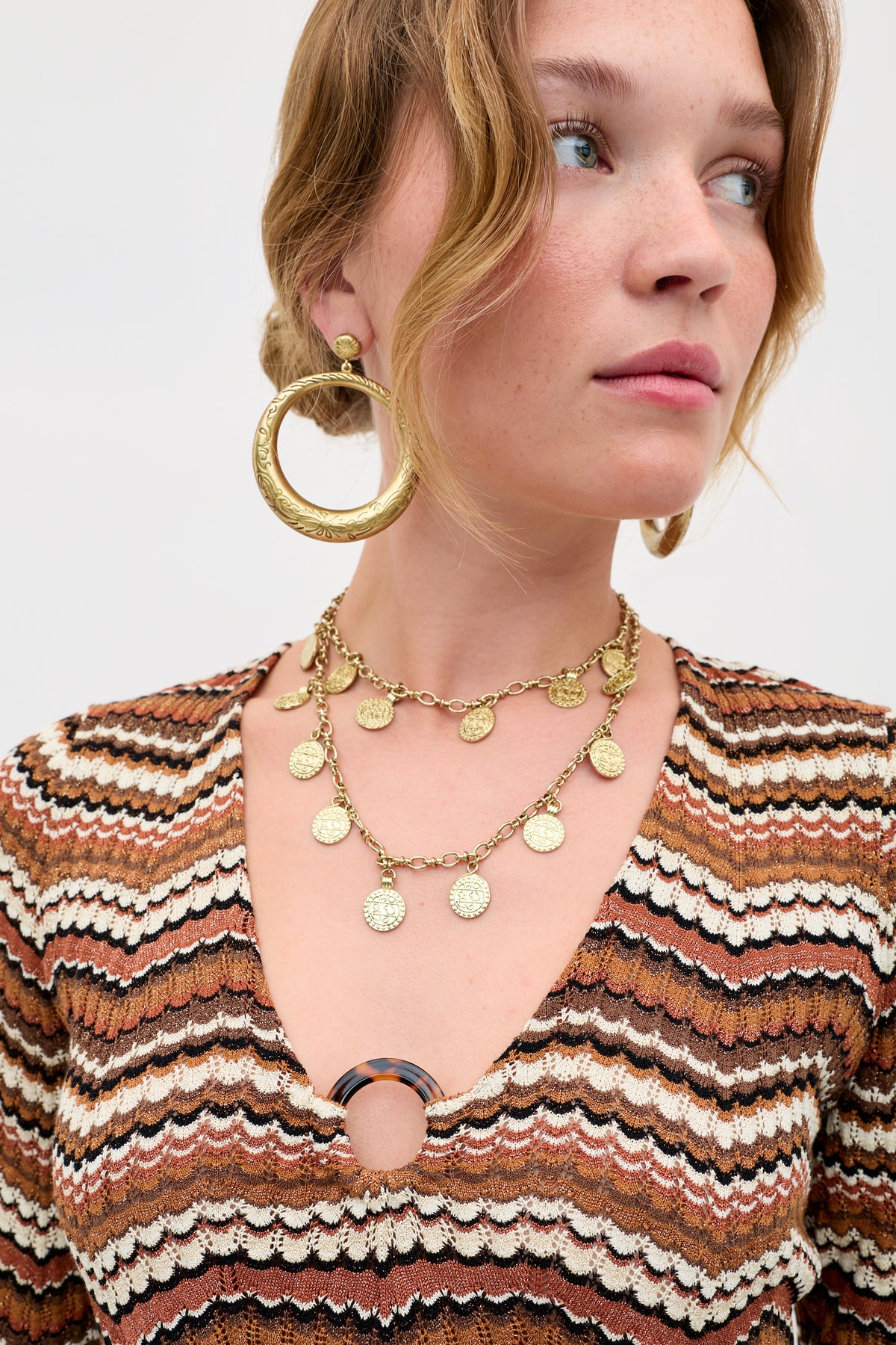 A woman wearing large gold hoop earrings, a layered gold coin necklace, and the Ivia - Maria Chevron Brown top with a zigzag pattern and circular clasp at the neckline.