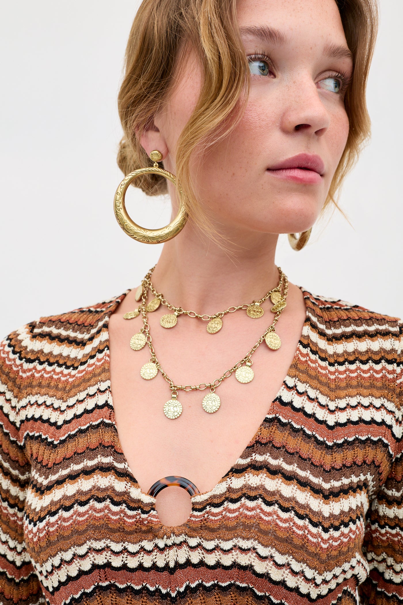 A woman wearing large gold hoop earrings, a layered gold coin necklace, and the Ivia - Maria Chevron Brown top with a zigzag pattern and circular clasp at the neckline.
