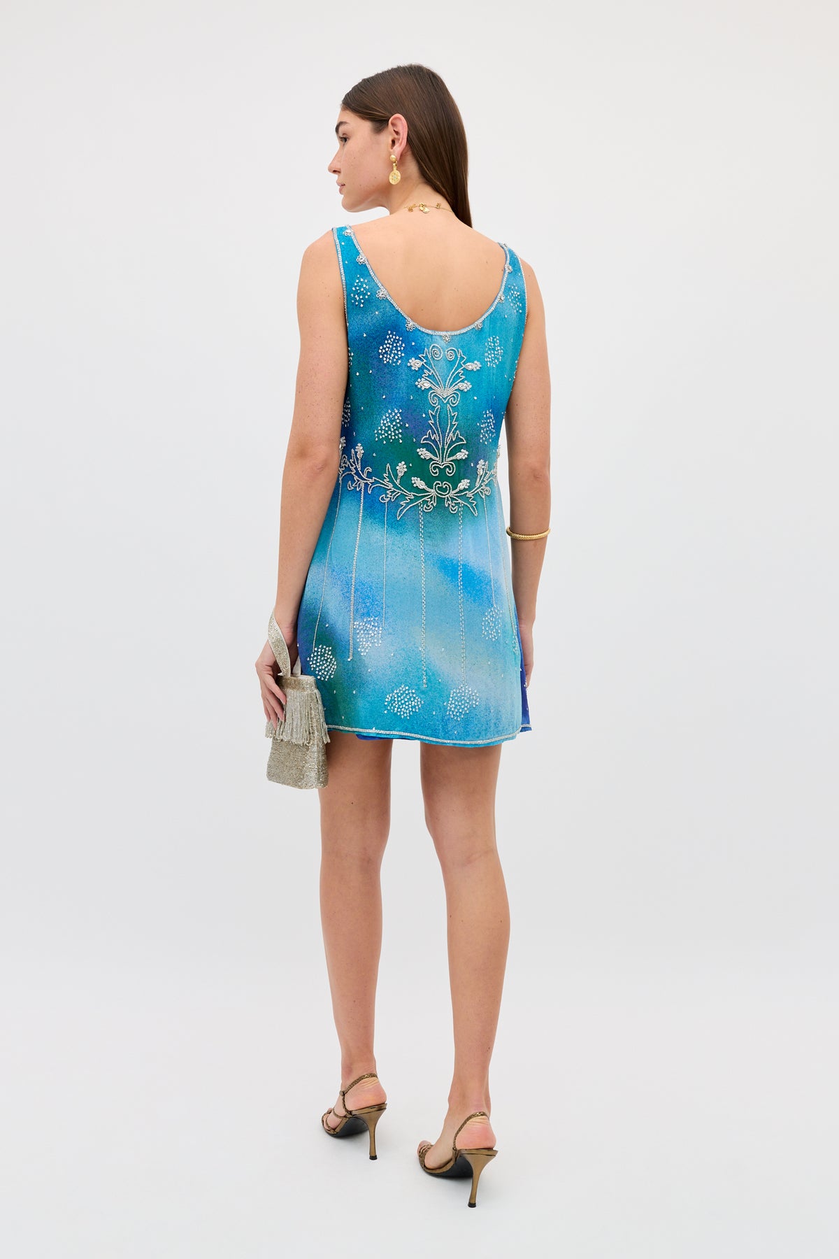 A woman stands facing away in a sleeveless Kaylor - Jessica Alexandra Blue mini dress with white floral designs. She holds a silver purse, wears black heels, gold earrings, and a bracelet. The background is plain and light-colored.