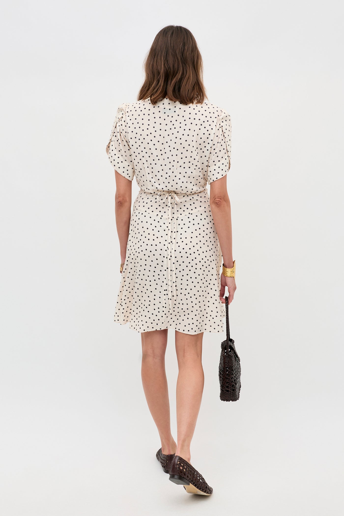A woman faces away, wearing the Sakura - Layla Dot Ivory short-sleeved, knee-length cream dress with black polka dots. She carries a dark woven handbag, wears flat shoes, her brown hair loose, and a gold bracelet on her wrist.