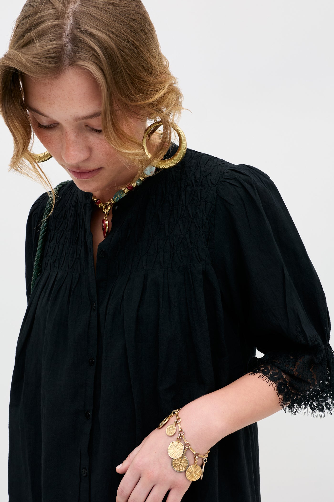 A woman wears the Siera - Black blouse with lace-trimmed sleeves, paired with large gold hoop earrings, a beaded necklace, and a coin-accented bracelet against a plain light background.