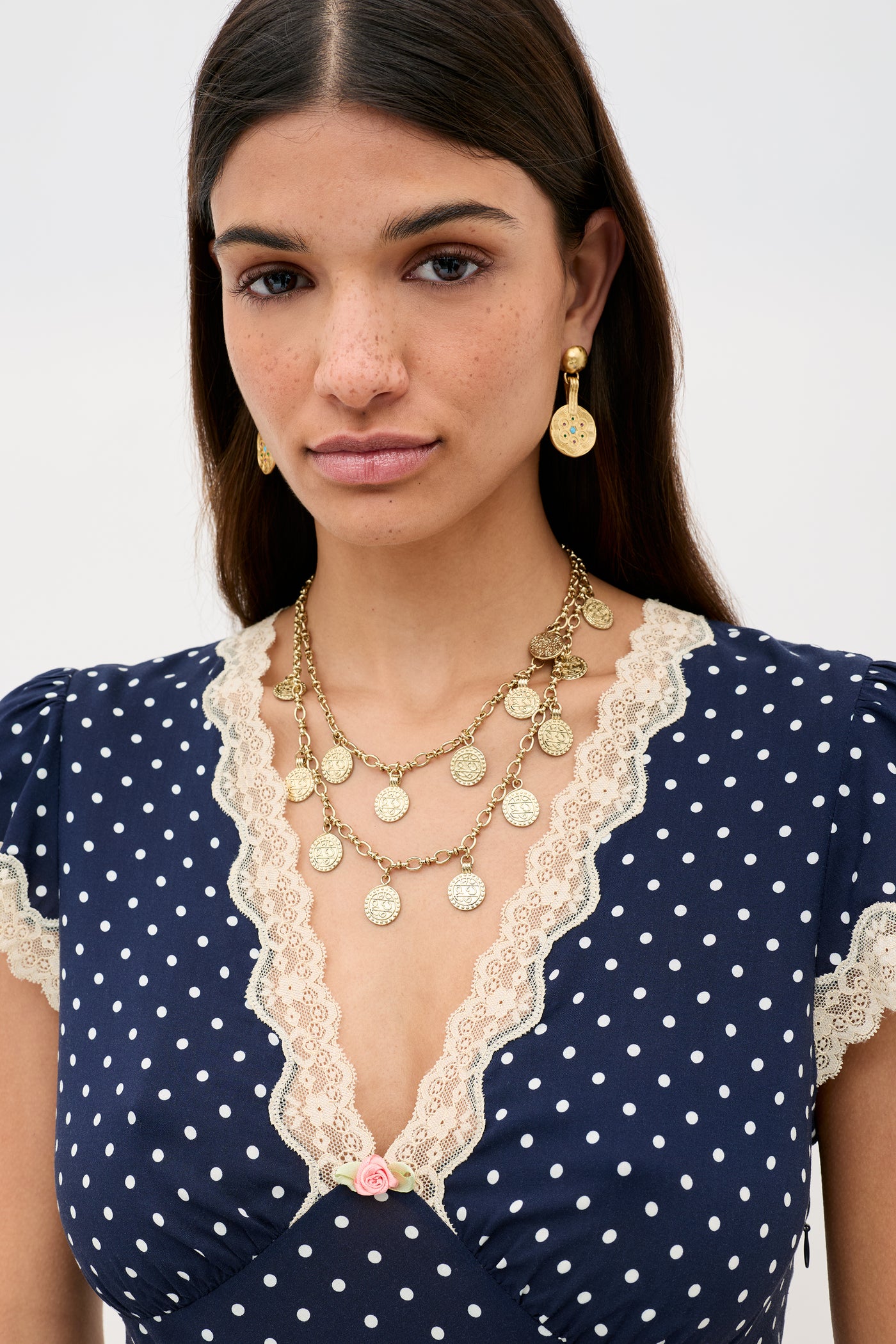 A woman with long dark hair wears the Clarice - Navy Polka Dot dress, styled like a vintage nightdress with lace trim. She accessorizes with gold coin earrings and a layered necklace, looking calmly at the camera.