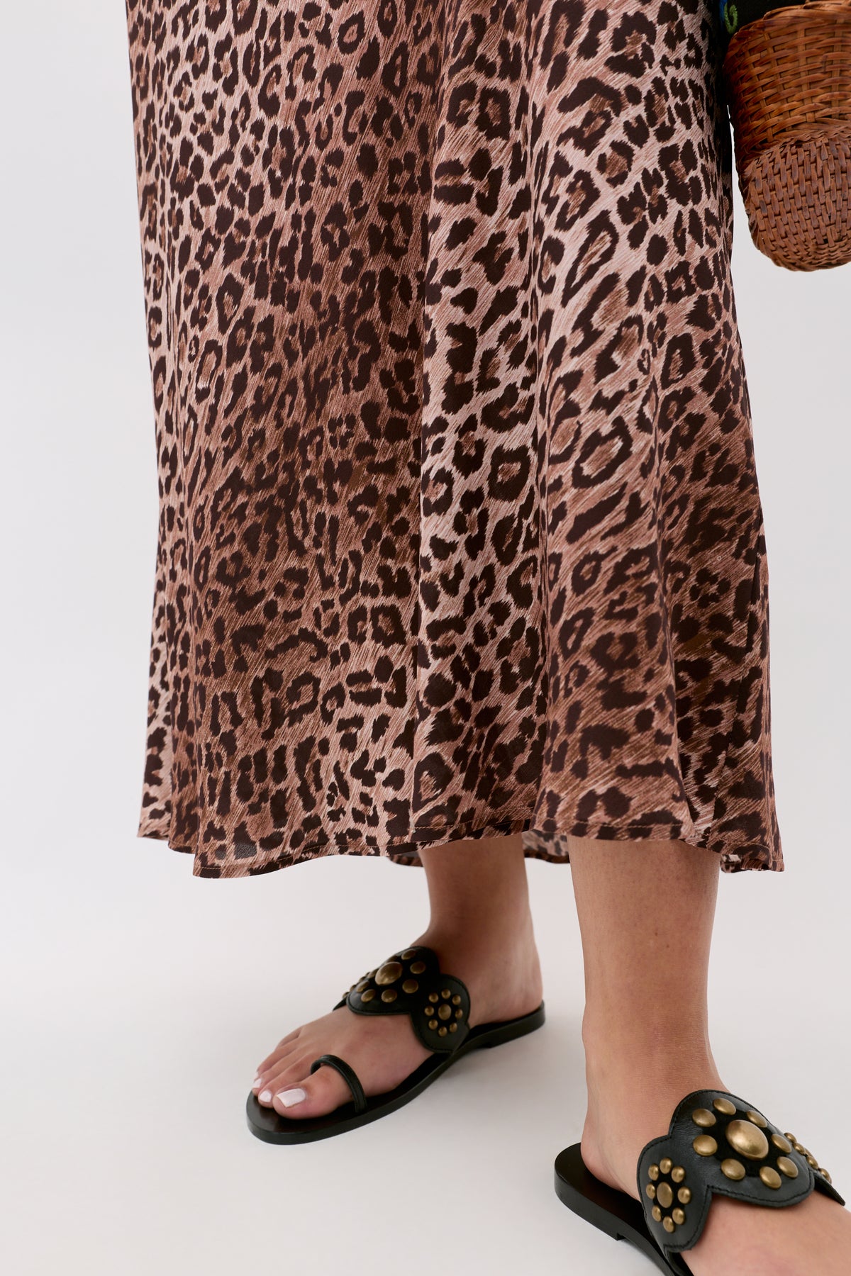 A person wearing the Kelly - Leopard Brown skirt, crafted from silk crepe de chine, and black sandals with gold accents stands on a light surface, holding a woven basket bag.