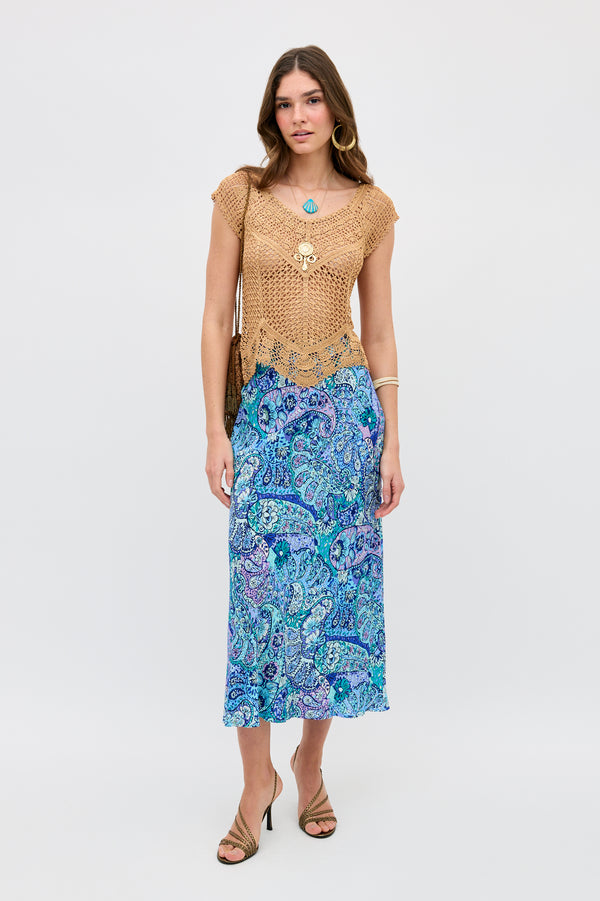 A woman faces the camera wearing the Kelly - Paisley Floral Blue skirt paired with a tan crocheted top. She accessorizes with sandals, jewelry, a brown bag, and has long brown hair. The background is white.