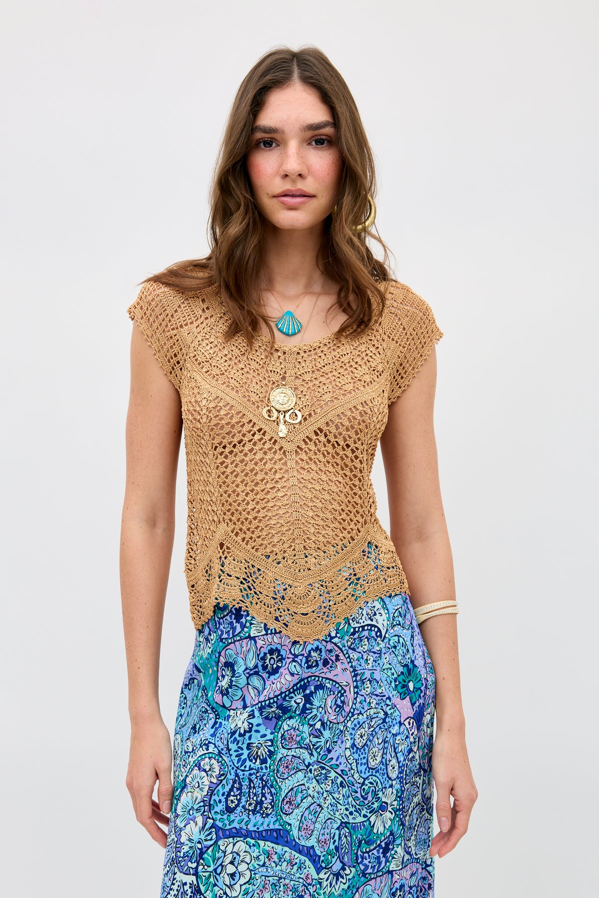 A woman with wavy brown hair wears the Kimbra - Gold necklace layered with other pieces, paired with a tan crochet top and a blue patterned skirt. She stands against a plain light background, looking at the camera with a neutral expression.