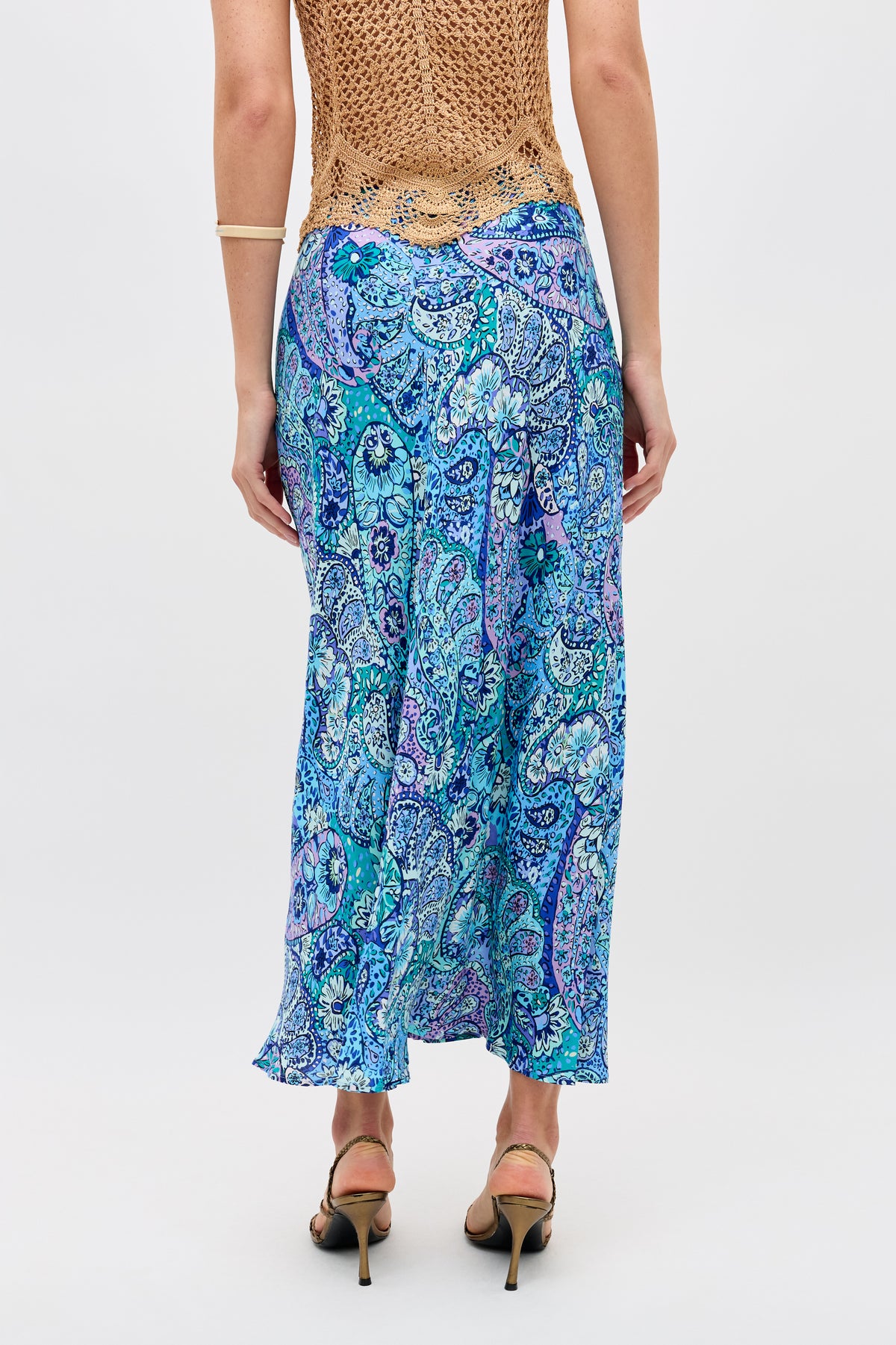 A person wearing a tan crochet top and the “Kelly - Paisley Floral Blue” long skirt stands facing away. They have a bracelet on one arm and are wearing black-heeled sandals. The background is plain white.