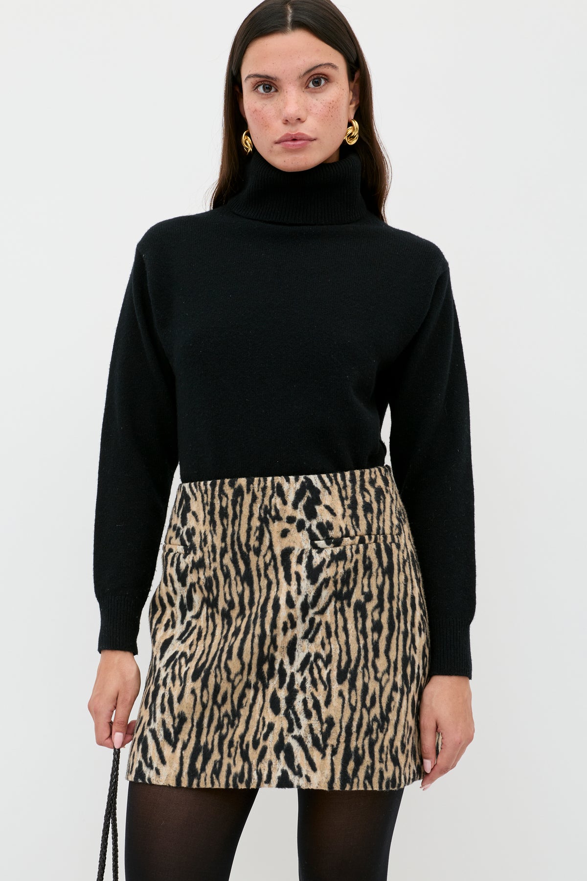 A woman in a black turtleneck, black tights, gold earrings, and the Mariah - Bohemia Leopard Brown mini skirt stands against a white background, holding a black purse—serving up chic noughties style.