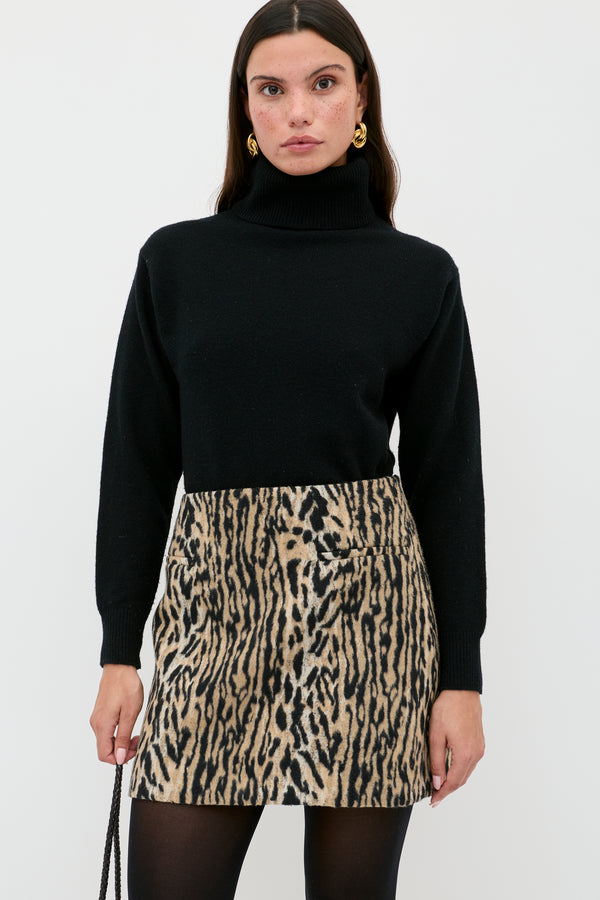 A woman in a black turtleneck, black tights, gold earrings, and the Mariah - Bohemia Leopard Brown mini skirt stands against a white background, holding a black purse—serving up chic noughties style.