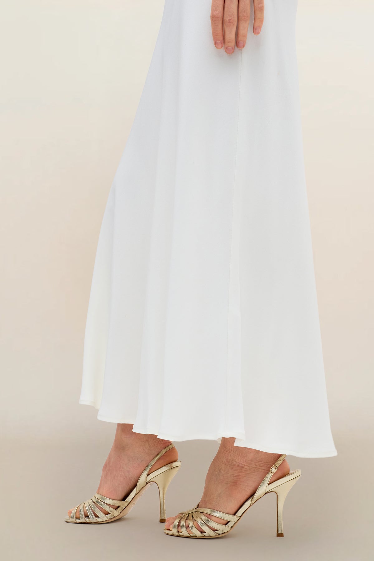 A person, reminiscent of a bridal specialist, wears the Joan - Ivory long white dress styled like a satin-back silk maxi skirt and gold high-heeled sandals, their hand gently resting on the dress, standing on a light-colored surface.