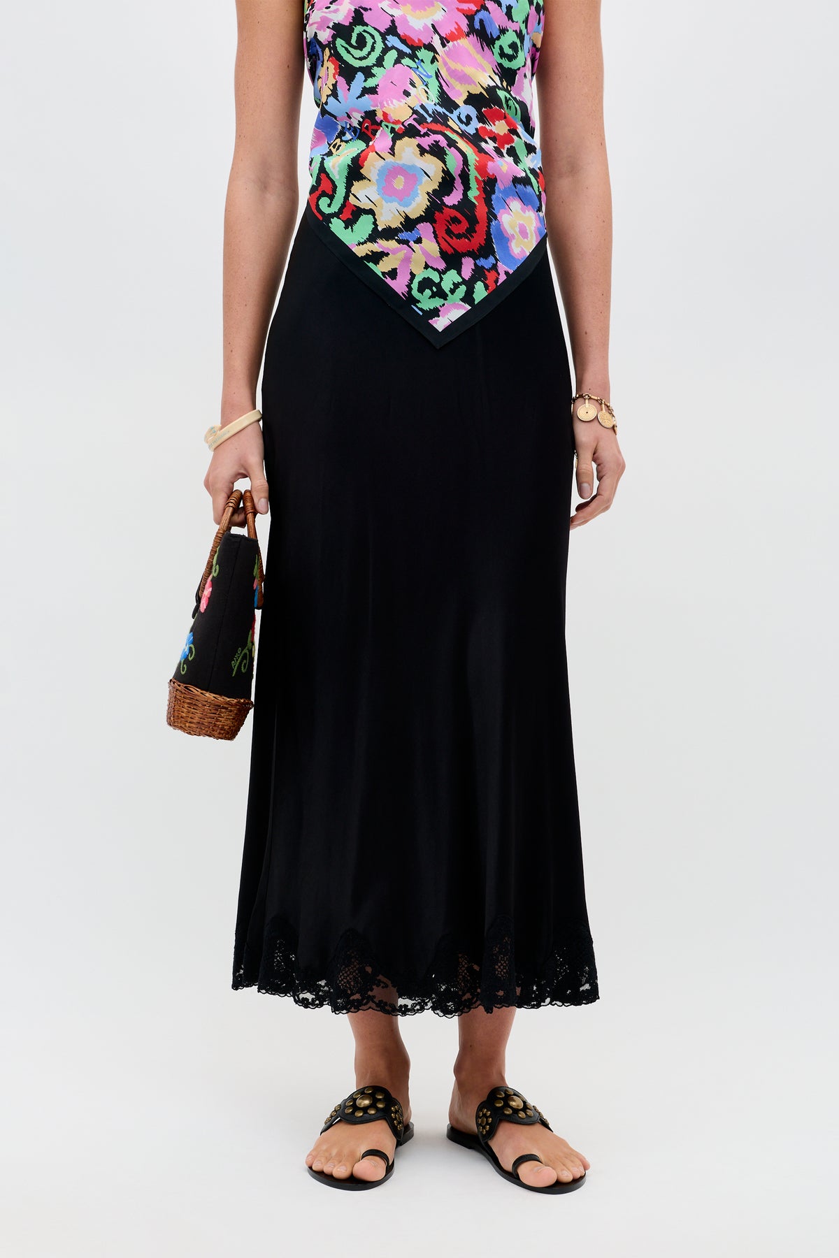 A woman wearing a colorful floral top and a black satin midi skirt with a lace hem, holding a woven handbag, and wearing the Crystal - Black sandals featuring gold details. The background is plain white.