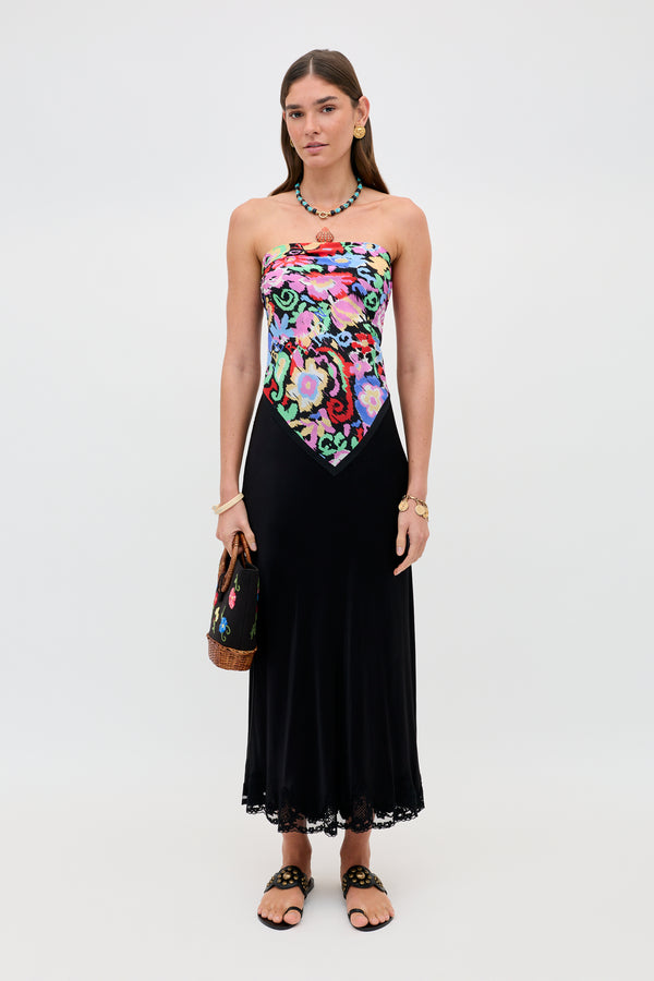 A woman wears the Crystal - Black dress, which features a colorful floral bodice and a flowing black satin midi skirt with delicate lace trim. She accessorizes with sandals, a necklace, a bracelet, and a handbag while standing against a plain white background.