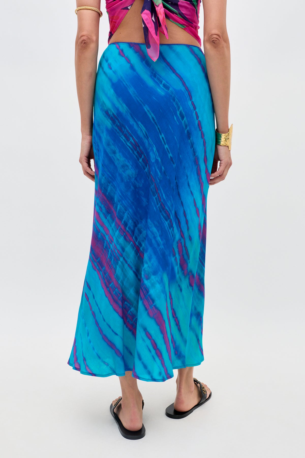 A person stands facing away, wearing the Ardith - Tie Dye Blue maxi skirt, a pink and purple top, black sandals, and gold jewelry, against a plain white background.
