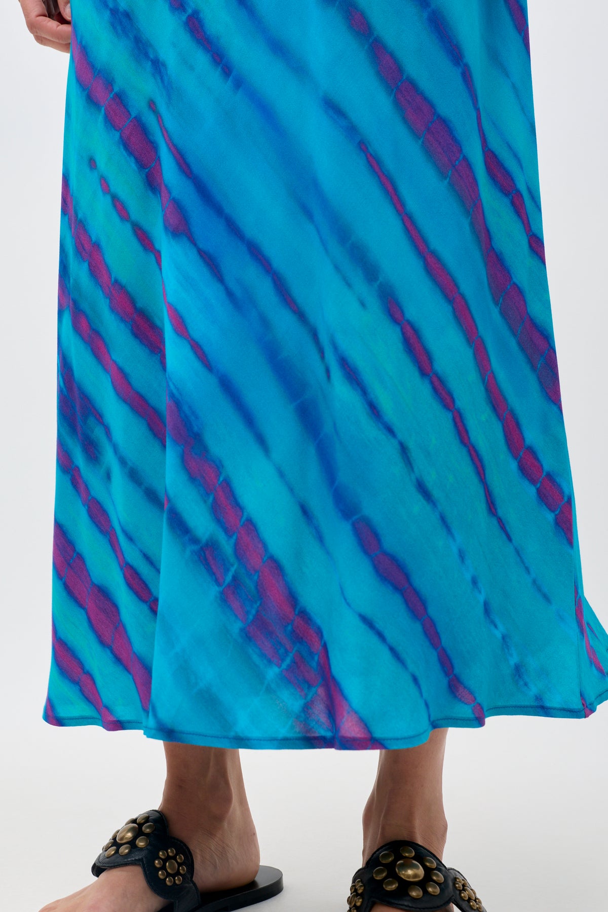 A person wearing the Ardith - Tie Dye Blue maxi skirt and black slide sandals with gold studs stands on a white background.