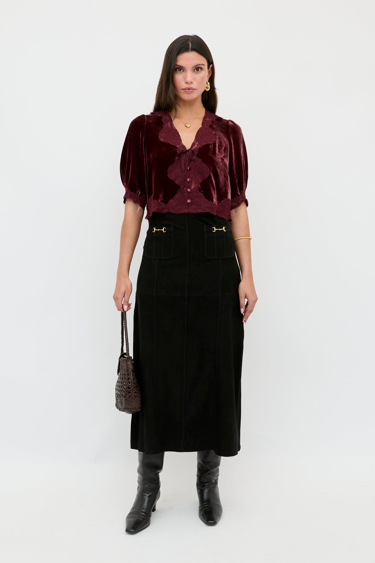 A woman with straight brown hair and a serious expression stands against a white background wearing the Edwina - Black high-waisted midi skirt with gold accents, a burgundy velvet blouse, black knee-high boots, and holding a small dark handbag.