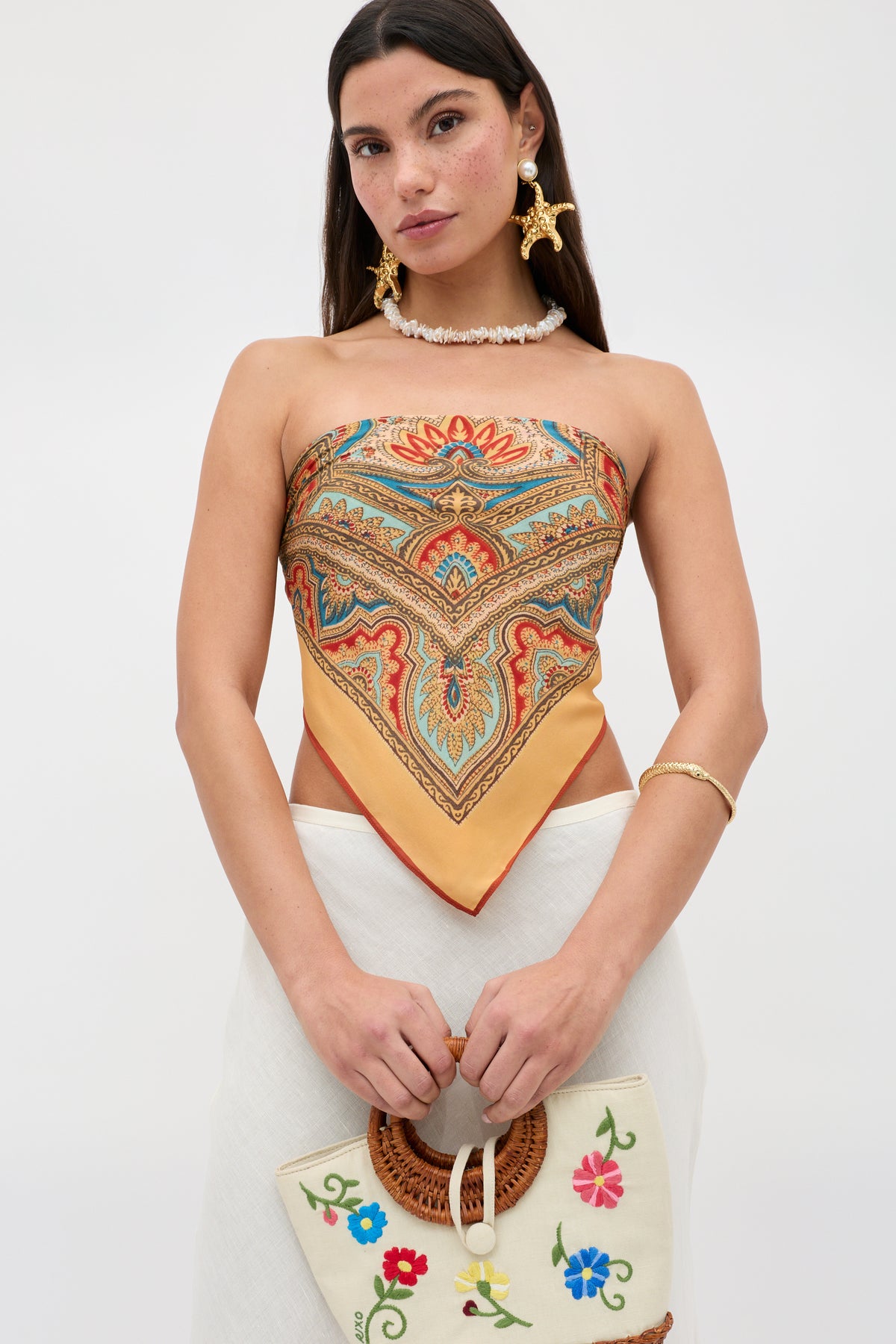 A woman wears the Menno - Paisley Golden patterned scarf top paired with a white skirt, holding an embroidered bag with floral designs. She accessorizes with statement earrings and a shell necklace, standing against a plain background.