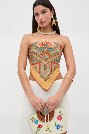 A woman wears the Menno - Paisley Golden patterned scarf top paired with a white skirt, holding an embroidered bag with floral designs. She accessorizes with statement earrings and a shell necklace, standing against a plain background.