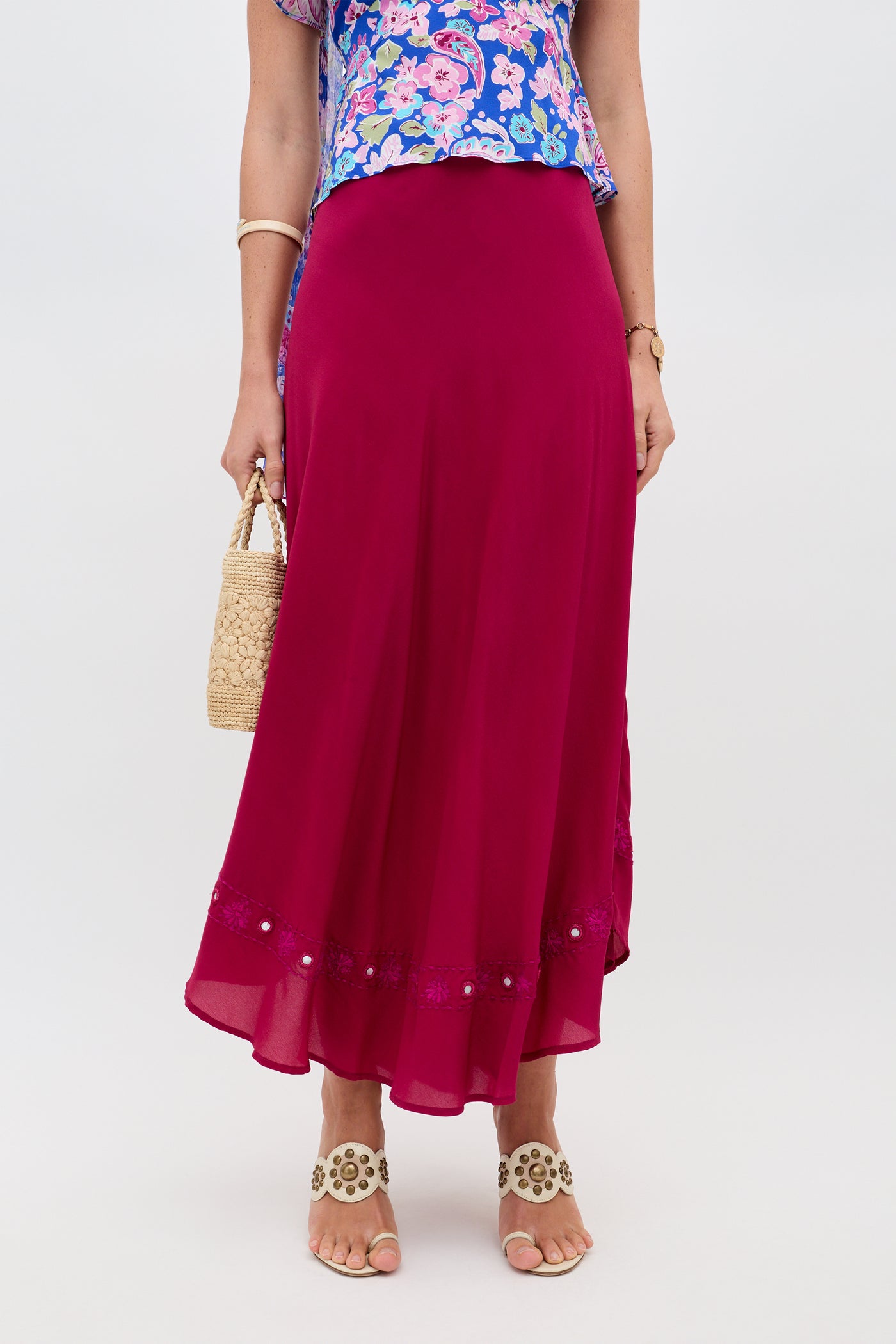 A person is wearing the Rudy - Mirror Embellishment Magenta Pink maxi skirt, paired with a floral top, beige sandals, and holding a woven handbag. The background is plain and light-colored.