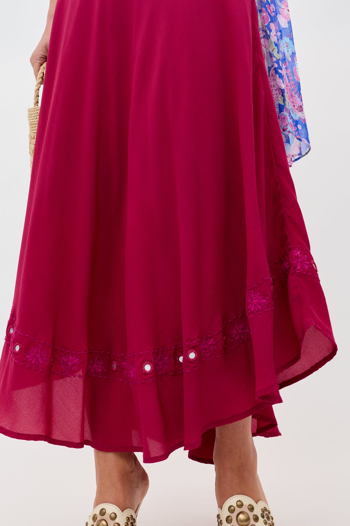 A person wearing the Rudy - Mirror Embellishment Magenta Pink skirt, featuring a flowing silhouette and floral trim, paired with white studded shoes; they hold a straw clutch and have a colorful scarf draped over their arm.