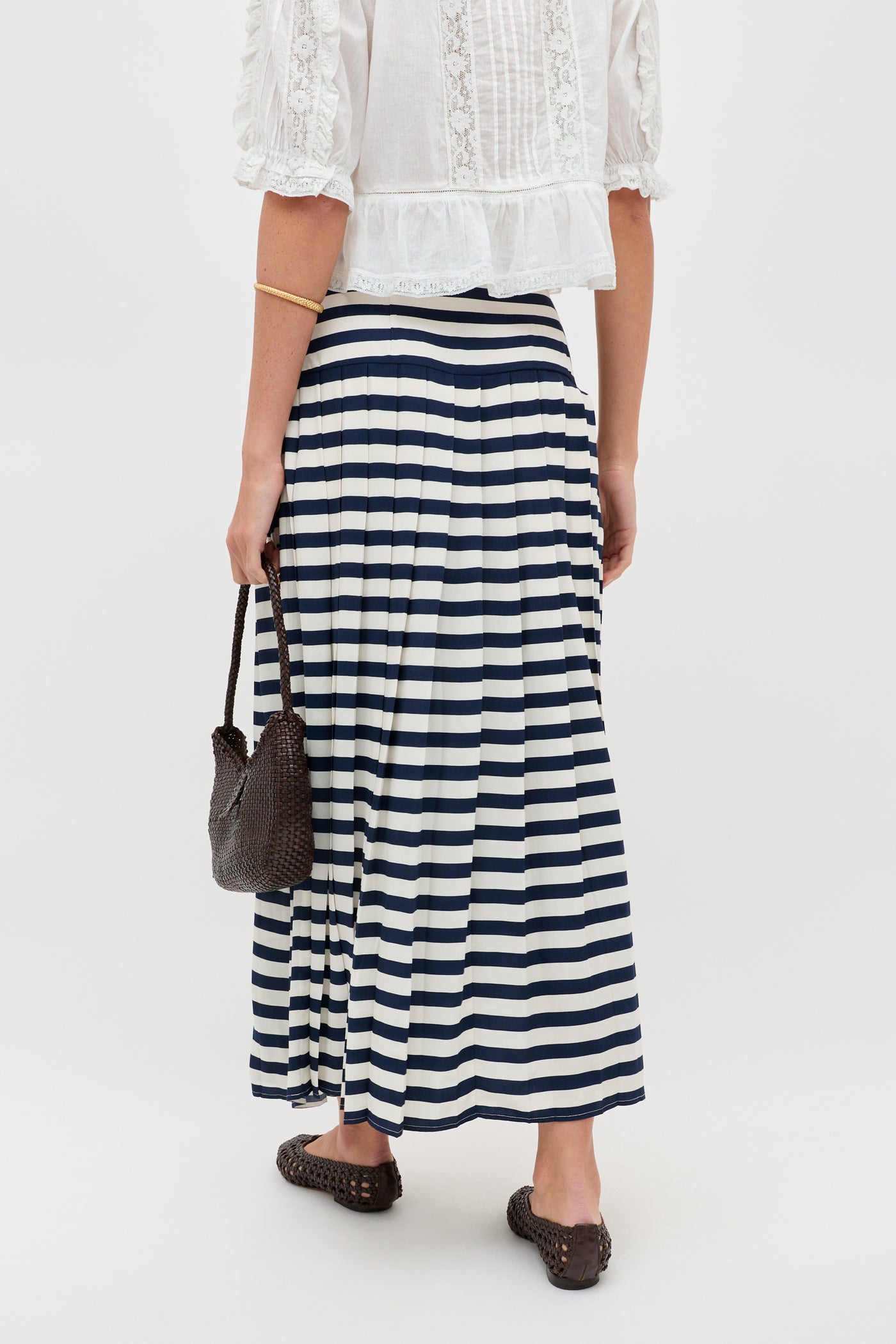 A person faces away, wearing the Una - Lea Stripe Navy pleated skirt with a white lace blouse. They hold a dark woven bag and wear dark woven flats. The background is plain and light.