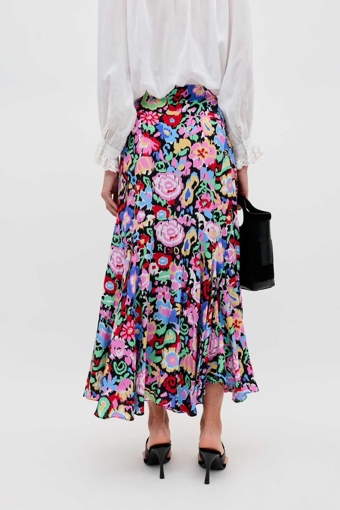 A person stands facing away, wearing the Libbie - Ikat Bloom Charcoal colorful floral midi skirt, a white long-sleeve blouse, black high-heeled sandals, and holding a small black bag against a plain background.