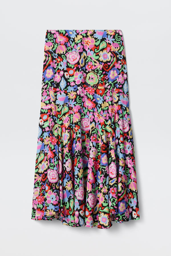 The Libbie - Ikat Bloom Charcoal skirt is knee-length with a vibrant multicolored floral pattern on a black background, featuring pink, red, purple, and green hues. It has a fitted waist and a flared, ruffled hem.