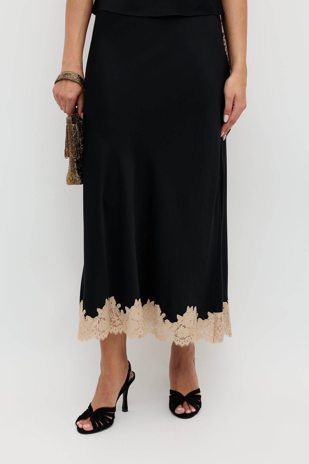 A woman wearing the Matilde - Black midi skirt with a beige lace hem, paired with black open-toe heels, gold bangles, and holding a gold beaded purse.