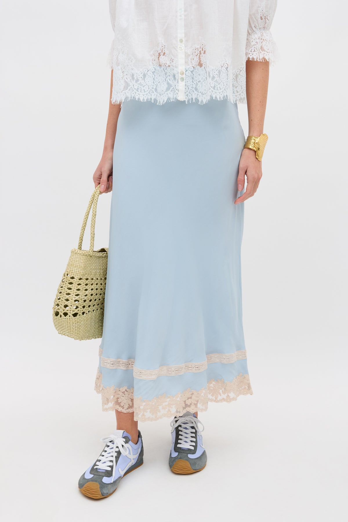 A person in the Costanza - Bluebell: a white lace blouse paired with a light blue skirt featuring lace trim, holding a woven straw bag and wearing running shoes. The image is cropped above the shoulders.