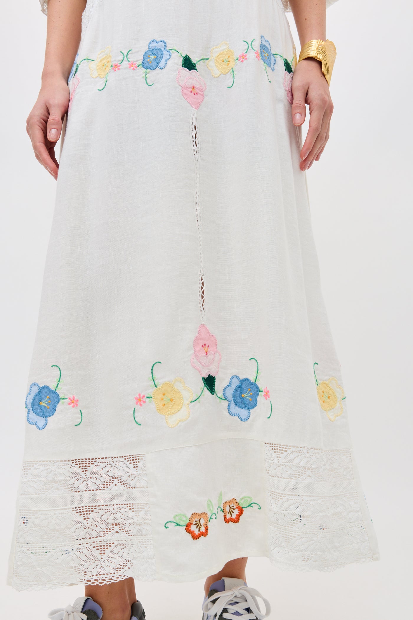 A person wearing the Paulina - Silje Embroidery Ivory skirt, featuring colorful floral embroidery near the hem and lace details, paired with gray sneakers and a gold bracelet.