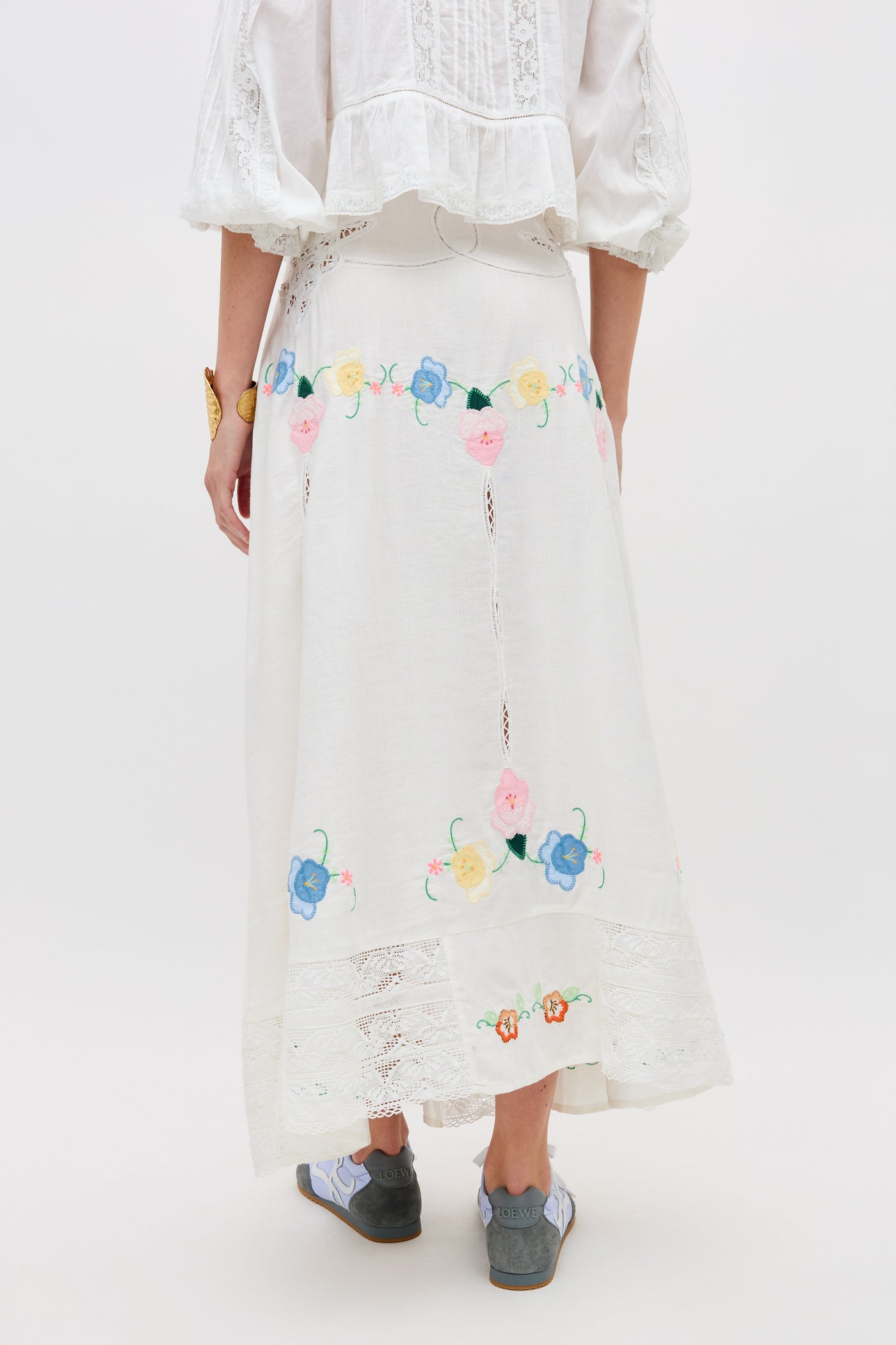 A person shown from the back wearing the Paulina - Silje Embroidery Ivory long white skirt with colorful floral embroidery and lace details, paired with a white blouse featuring puffed sleeves and gray shoes.