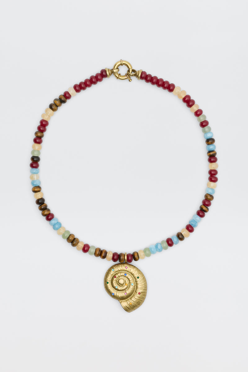 The Parma - Multi beaded necklace showcases red, blue, brown, and white beads with a gold spiral shell pendant adorned with small colored gemstones, elegantly displayed on a white background.