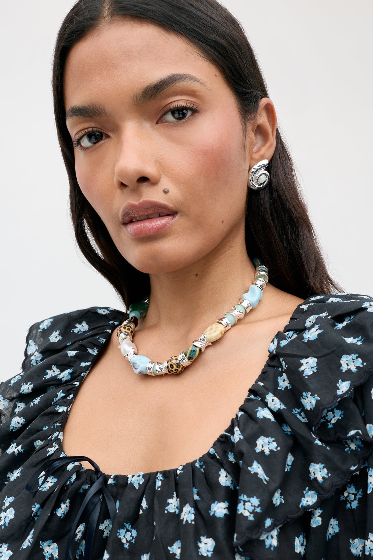 A woman with long dark hair wears the Maxina - Blue dress, styled with a colorful beaded necklace, round earrings, and natural makeup, looking confidently at the camera against a plain background.