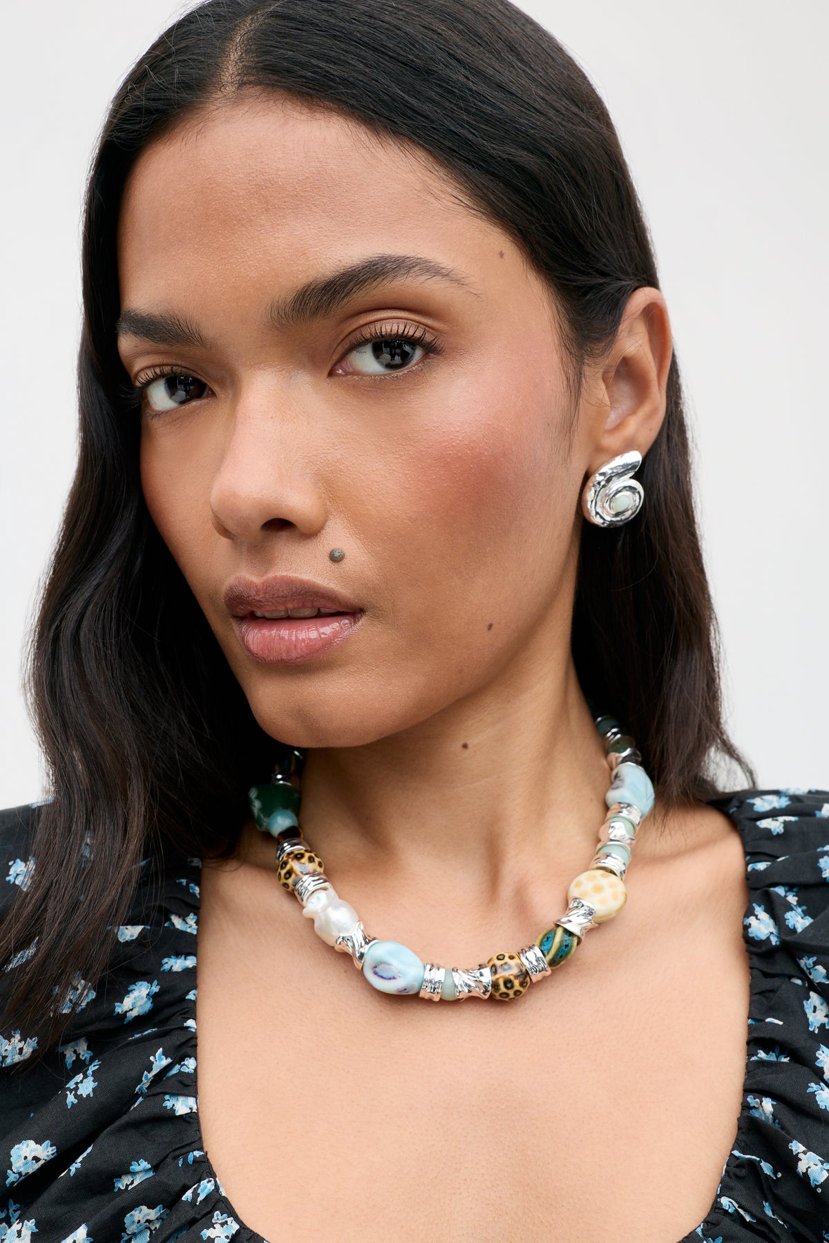 Woman with long dark hair, wearing a floral black top, silver statement earrings, and the Maxina - Blue chunky blue and white beaded necklace, looking directly at the camera with a neutral expression.