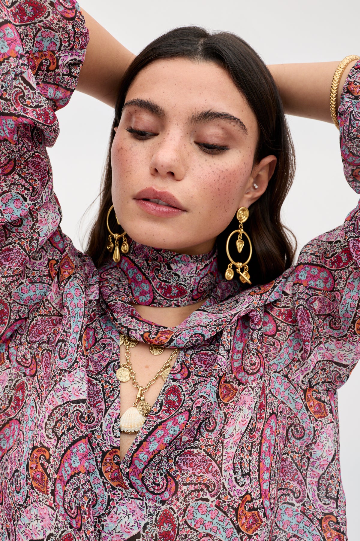 A woman with long brown hair and freckles wears the Moss - Bourton Red blouse, featuring a vintage-inspired paisley pattern. She accessorizes with gold jewelry and large earrings, her eyes closed and arms raised behind her head.