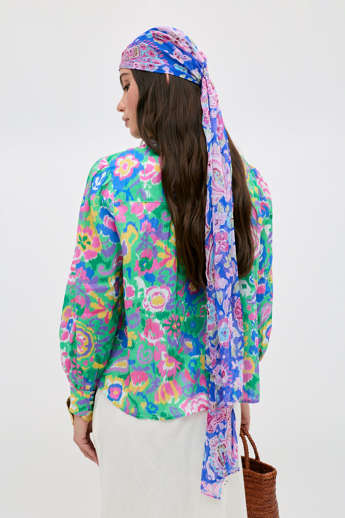 A woman facing away is wearing the Blake - Ikat Bloom Green blouse, a long purple and blue patterned headscarf, and a white skirt. She holds a woven bag in her left hand against a plain, light-colored background.