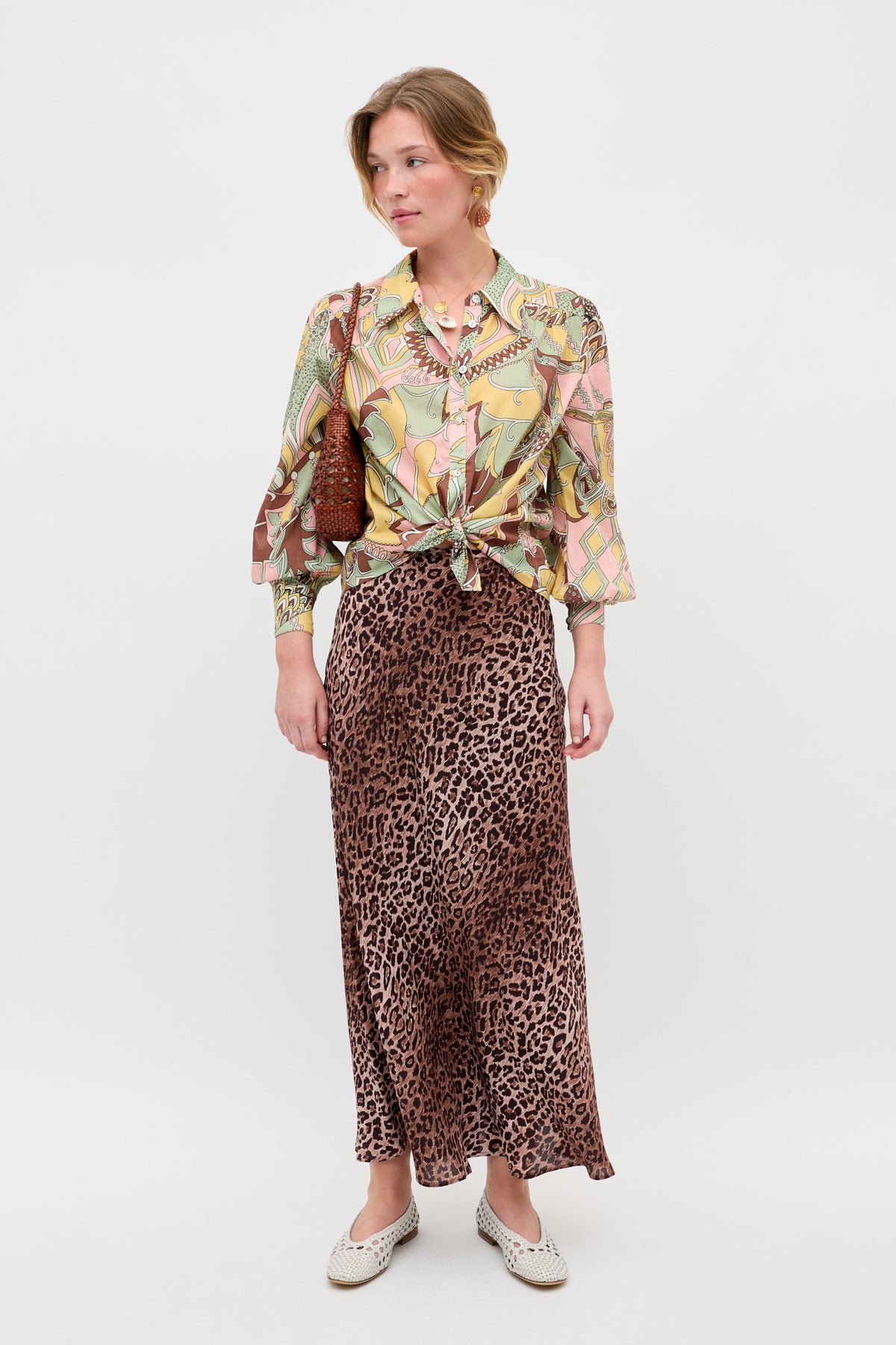 A woman stands against a plain background, wearing a colorful patterned blouse tied at the waist, the Kelly skirt in Leopard Brown—crafted from luxurious silk crepe de chine—a pair of white flat shoes, and a brown shoulder bag. She looks slightly to her left.