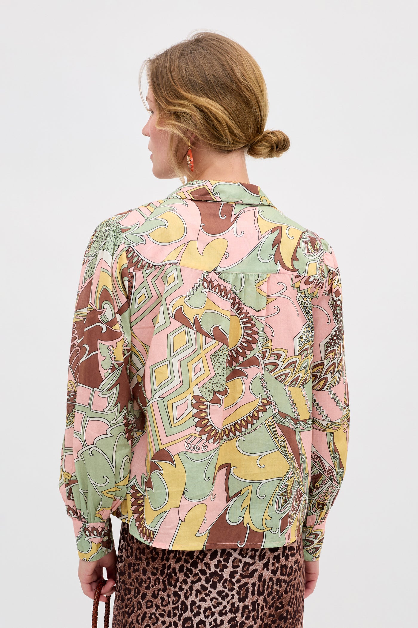 A woman with blonde hair in a low bun is seen from behind, wearing the Blake - Bianca Abstract Green blouse featuring pink, green, yellow, and brown abstract shapes, paired with a brown leopard print skirt.