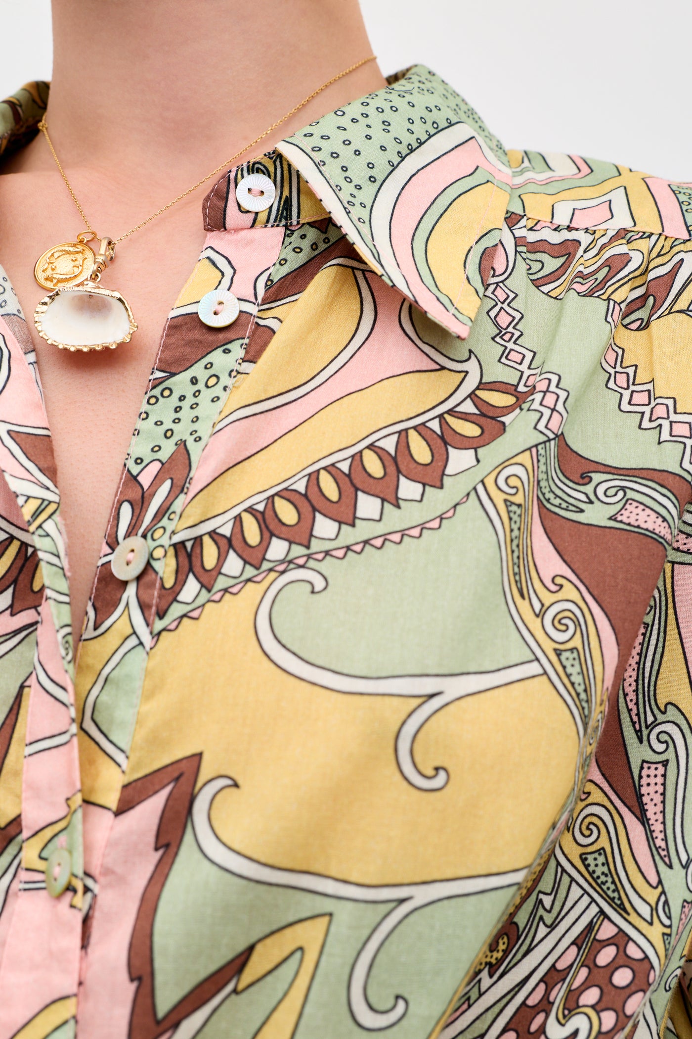 A close-up of a person wearing the Blake - Bianca Abstract Green shirt, featuring colorful swirling patterns in yellow, green, pink, and brown, paired with a gold necklace with a white shell pendant and round charm.