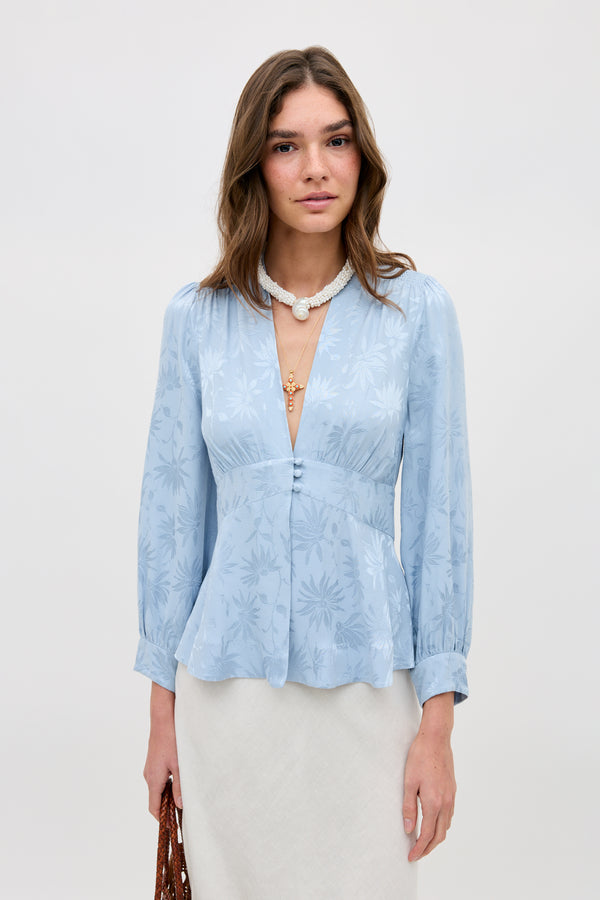 A woman stands against a plain background wearing the Lea - Daisy Jacquard Bluebell, a light blue, long-sleeve blouse with a floral pattern and deep V-neck. She holds a brown woven bag and accessorizes with a white necklace featuring a pendant.
