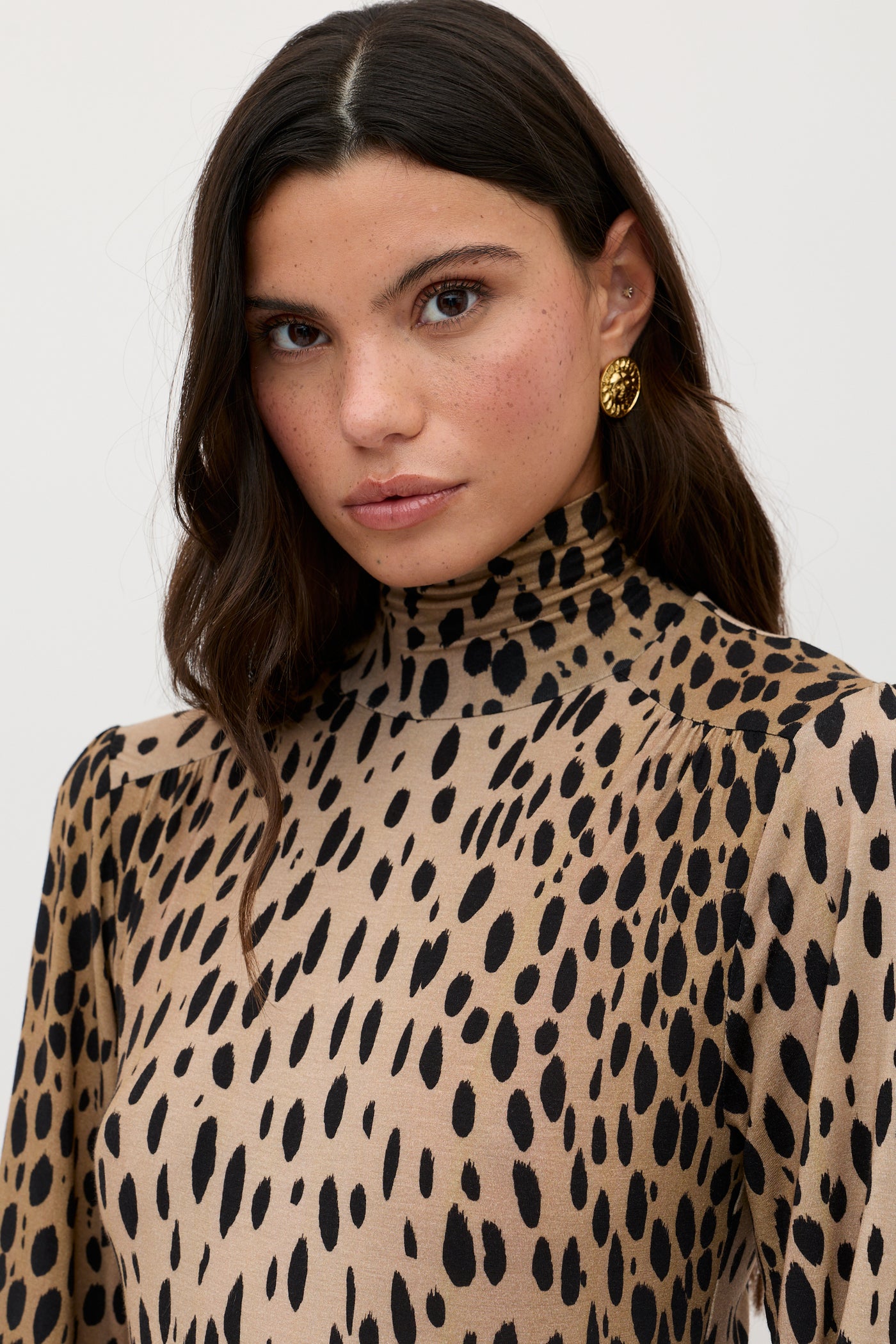 A woman with long dark hair wears the Suki - Antelope Spot Black high-neck, long-sleeve dress and gold round earrings, standing against a plain light background.