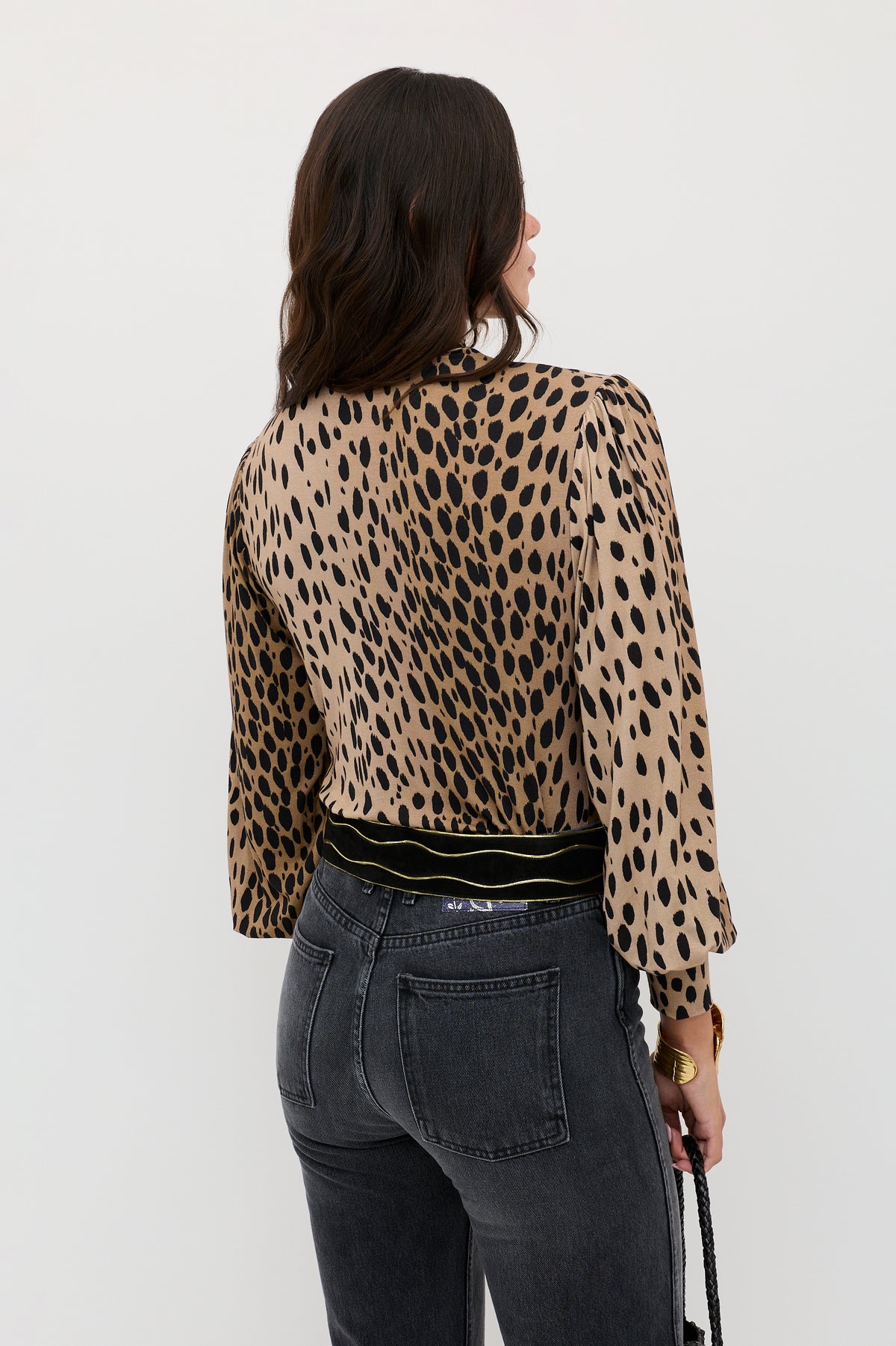 A woman with wavy dark hair, seen from behind, wears the long-sleeved Suki - Antelope Spot Black top and high-waisted black jeans, holding a black strap bag against a plain white background.
