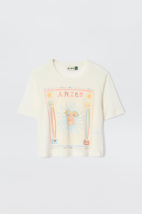 The Ria - Aries white short-sleeve T-shirt displays a vibrant Aries graphic with "ARIES" at the top, a central ram, sun rays, and pink, orange, and blue astrological symbols.