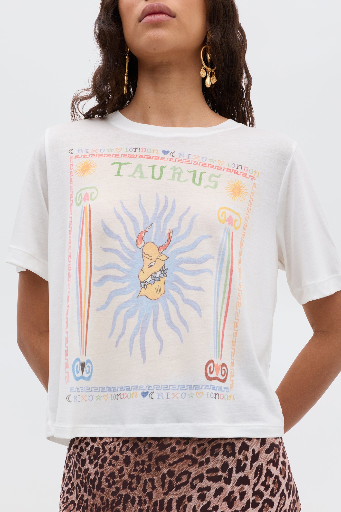 A woman wears the Ria - Taurus white t-shirt featuring a colorful Taurus bull design, paired with a leopard print RIXO skirt. She has long hair, gold earrings, and stands against a plain backdrop.